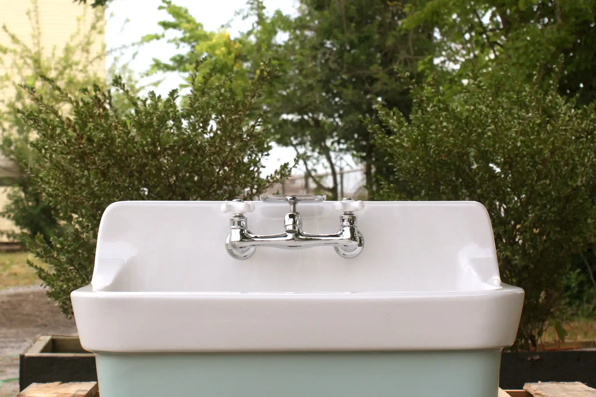A classic porcelain utility sink with a chrome faucet, placed outdoors against a backdrop of green shrubs. | ACE DECOR