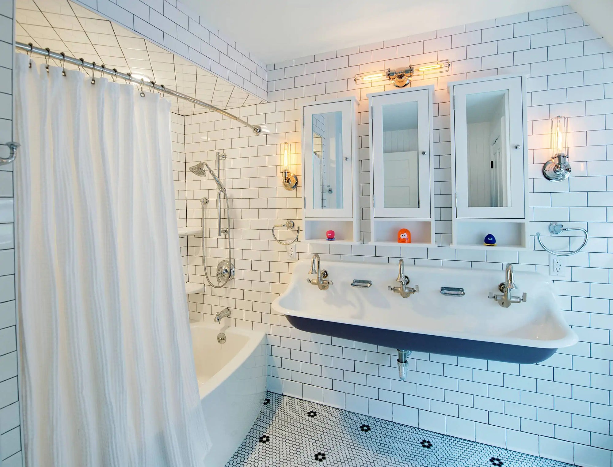 A bright bathroom with white subway tiles features a large vintage wall mount sink with multiple faucets beneath mirrored cabinets. |ACE DECOR