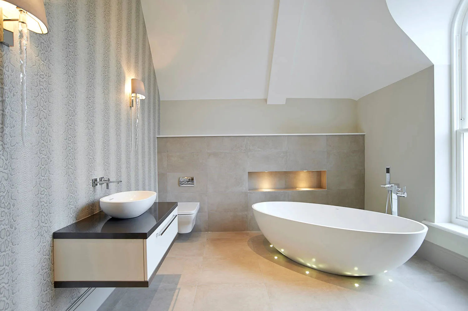 A modern style bathroom with a freestanding white bathtub, a stylish sink on a dark topped cabinet, and wall mounted lights