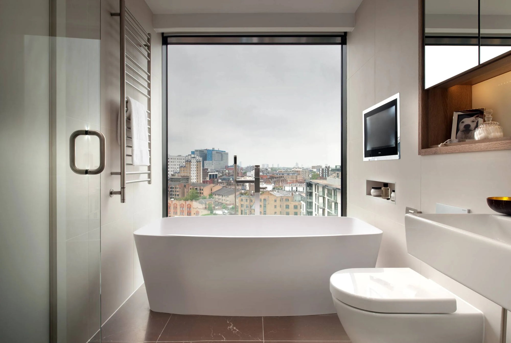 A modern bathroom featuring a city-view window, white freestanding bathtub, glass-enclosed shower, wall-mounted TV, and wooden shelf in soft neutral tones.