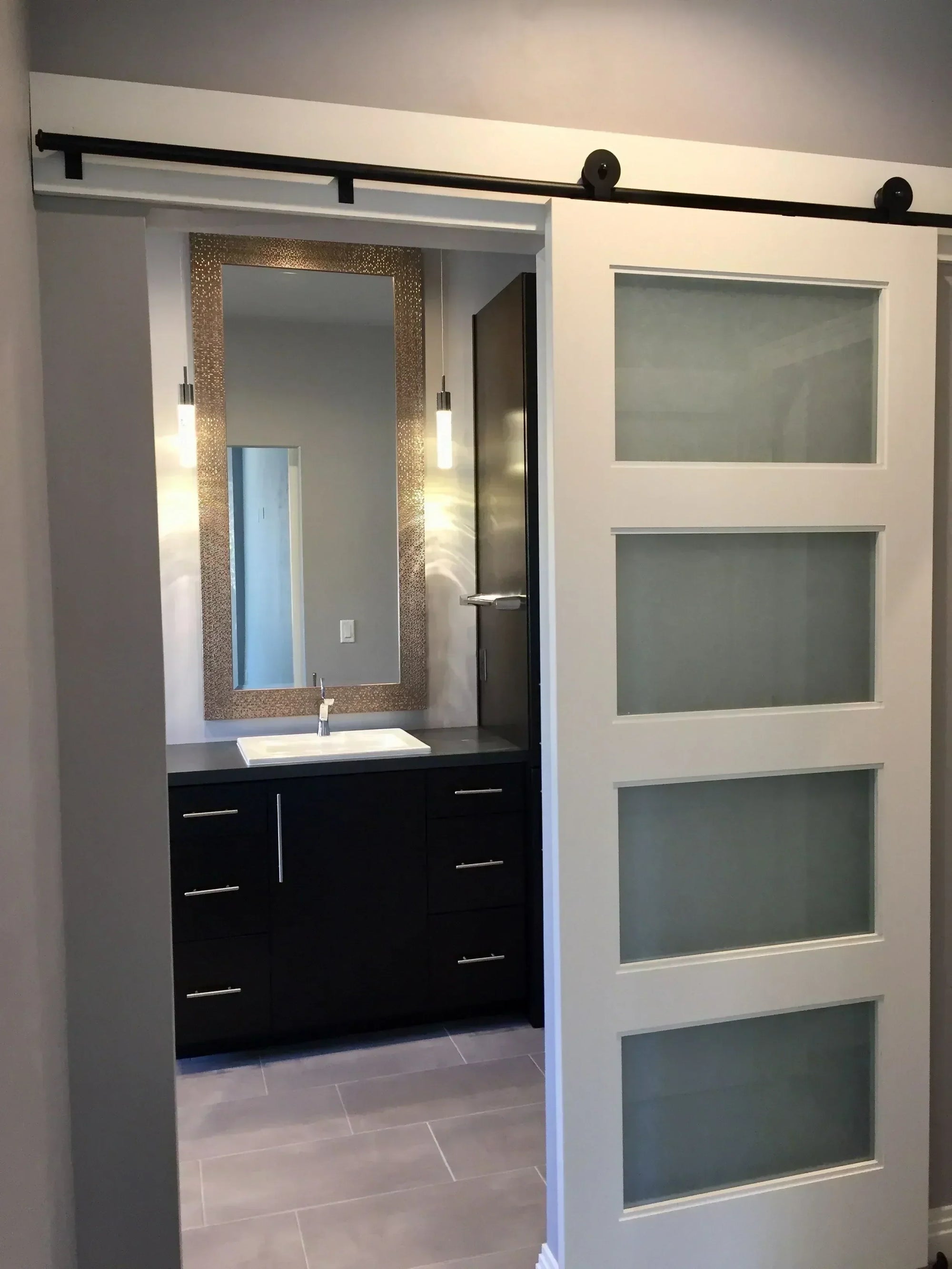 Modern sliding barn door with horizontal glass panels in a neutral-toned bathroom, complemented by a gold-accented mirror and dark cabinetry creating striking contrast against pale tile flooring, emphasizing minimalist contemporary design