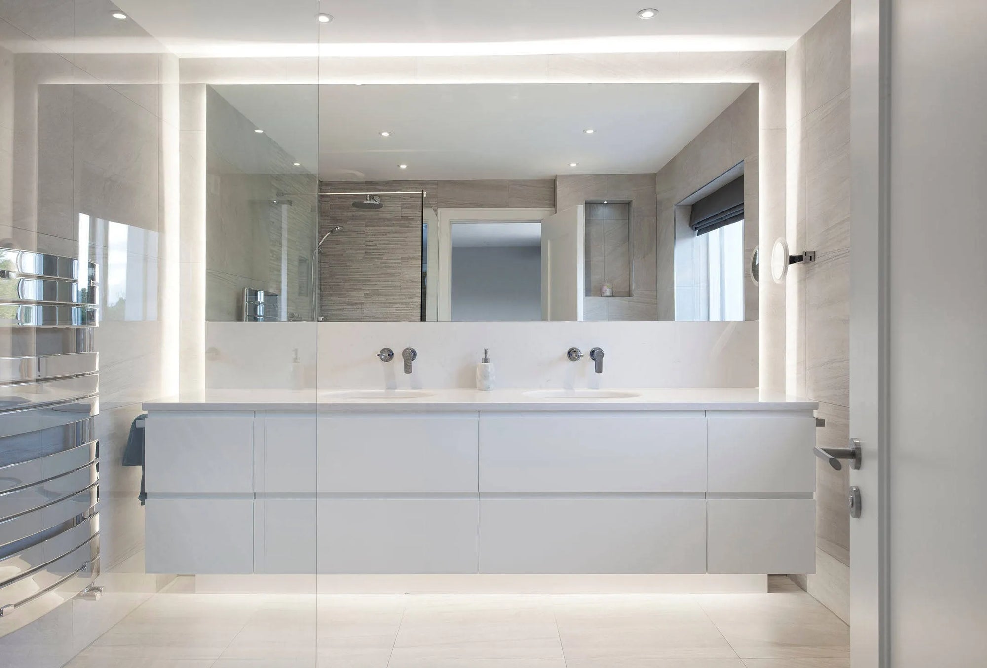 Modern minimalist bathroom with a sleek white double-sink vanity, large mirror reflecting windows/textured walls, soft ambient lighting, bright and clean aesthetic.