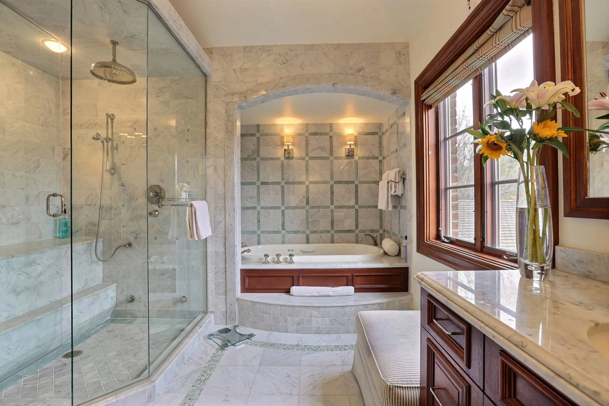 Luxurious bathroom with glass-enclosed shower, alcove bathtub, marble vanity with yellow-white flowers, neutral tones, warm wood accents, arched lighting.