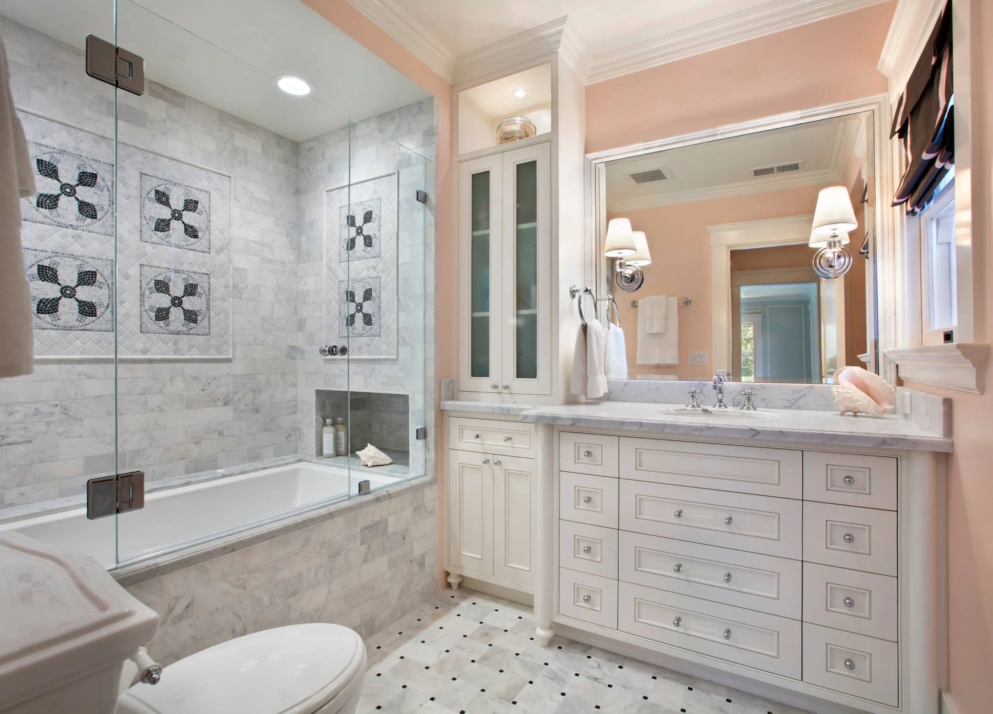A modern elegant bathroom featuring soft pink walls, white double vanity with mirrored cabinet, glass-enclosed bathtub, geometric floor tiles and decorative tiled shower walls.