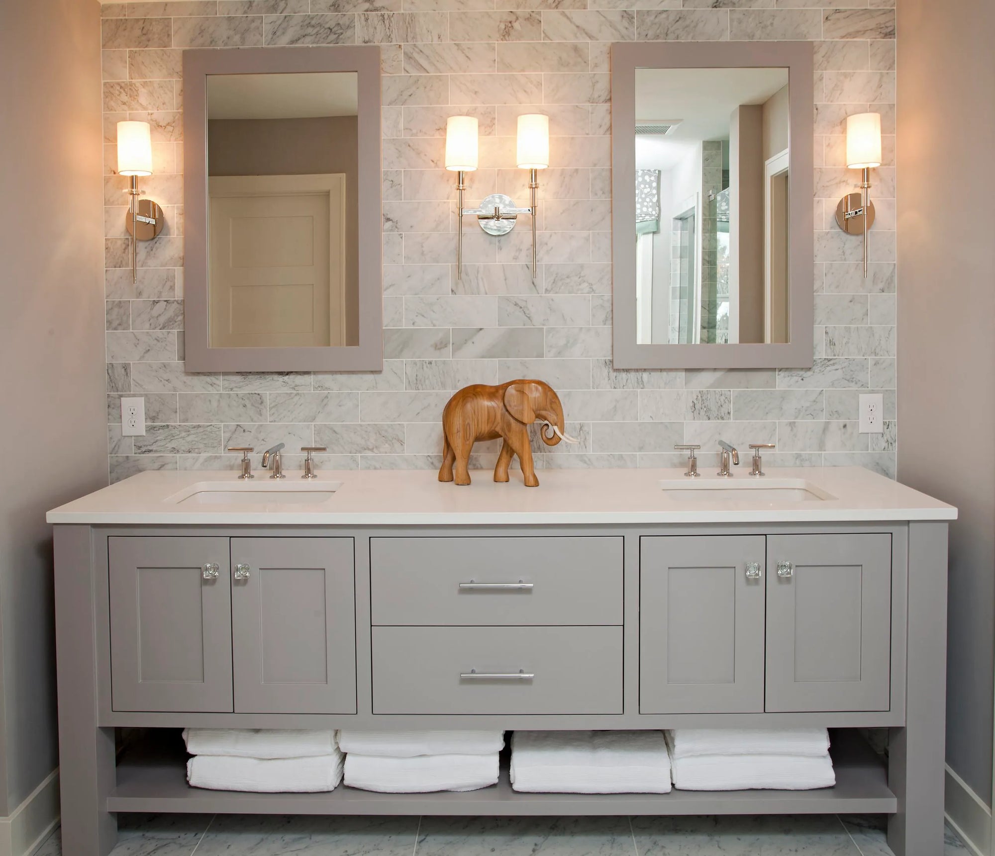 Modern minimalist bathroom with gray double-sink vanity, white marble countertop featuring an elephant figurine, two mirrored sconces with warm lighting.
