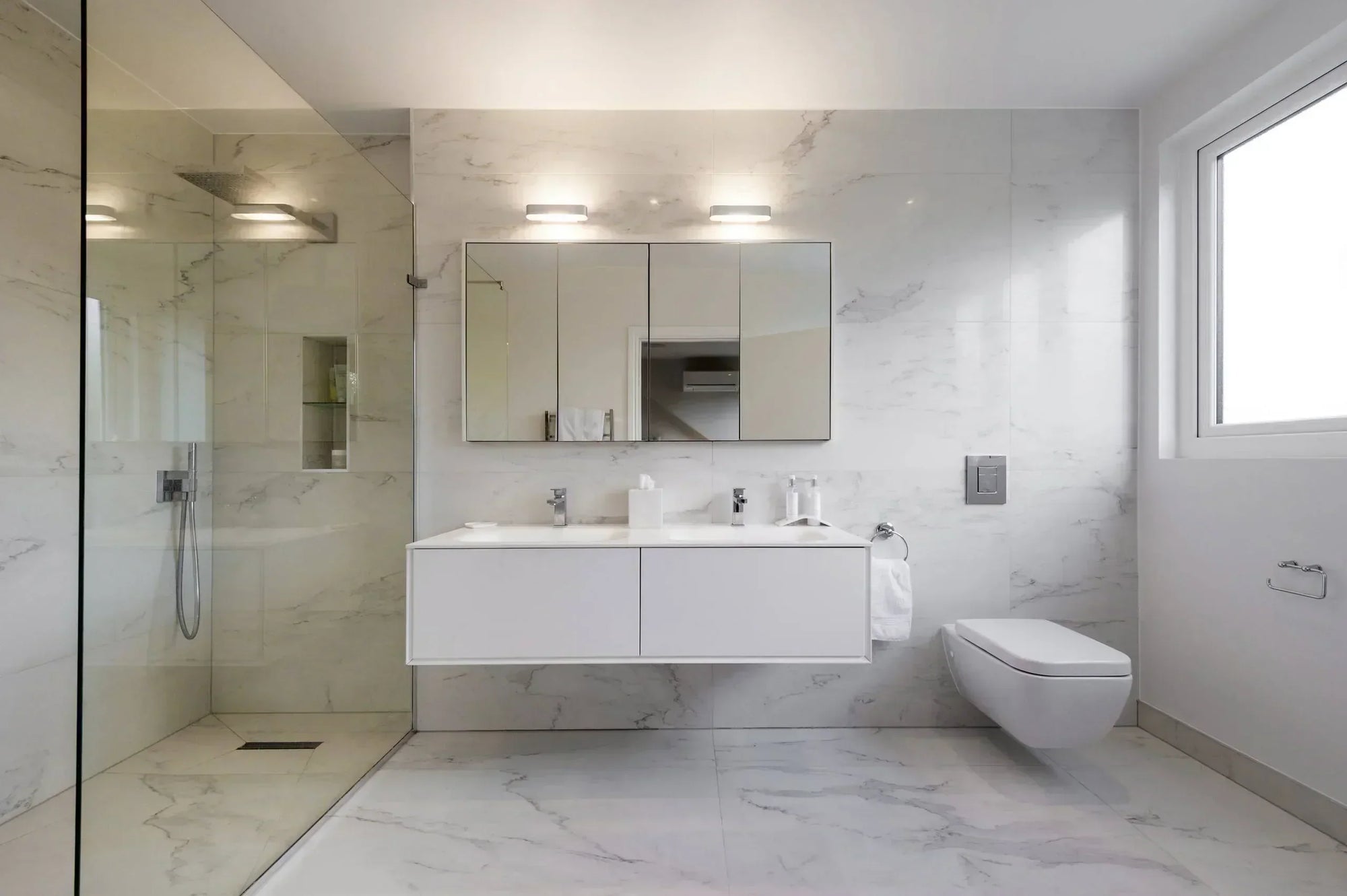A modern minimalist bathroom in white marble with a glass shower, floating vanity, wall-mounted toilet, and natural light window.