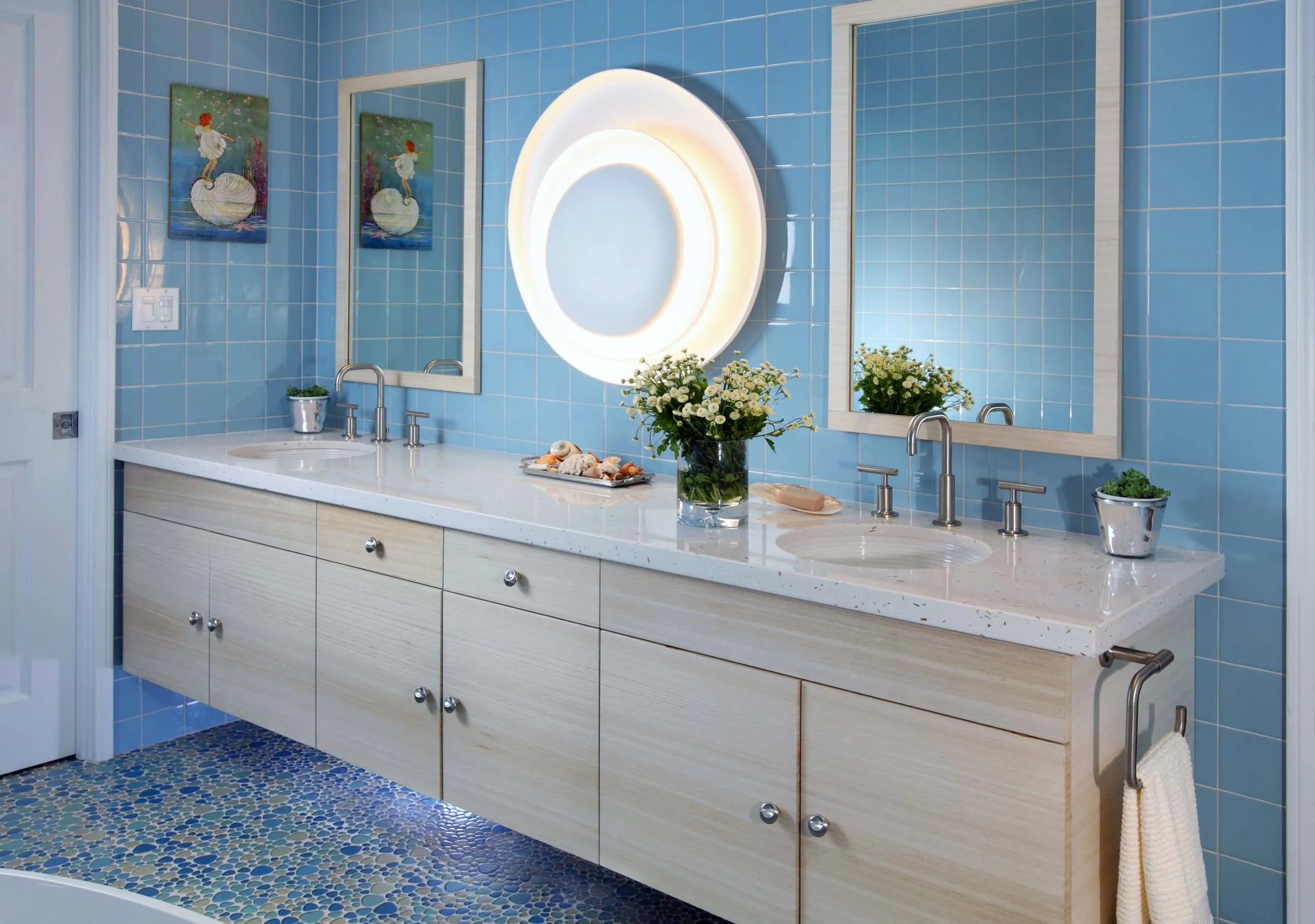 Modern bathroom with light-blue tiled walls, light-gray marble vanity with dual sinks, round mirror with ambient lighting, fresh floral arrangements, blue patterned floor tiles, and framed artwork.