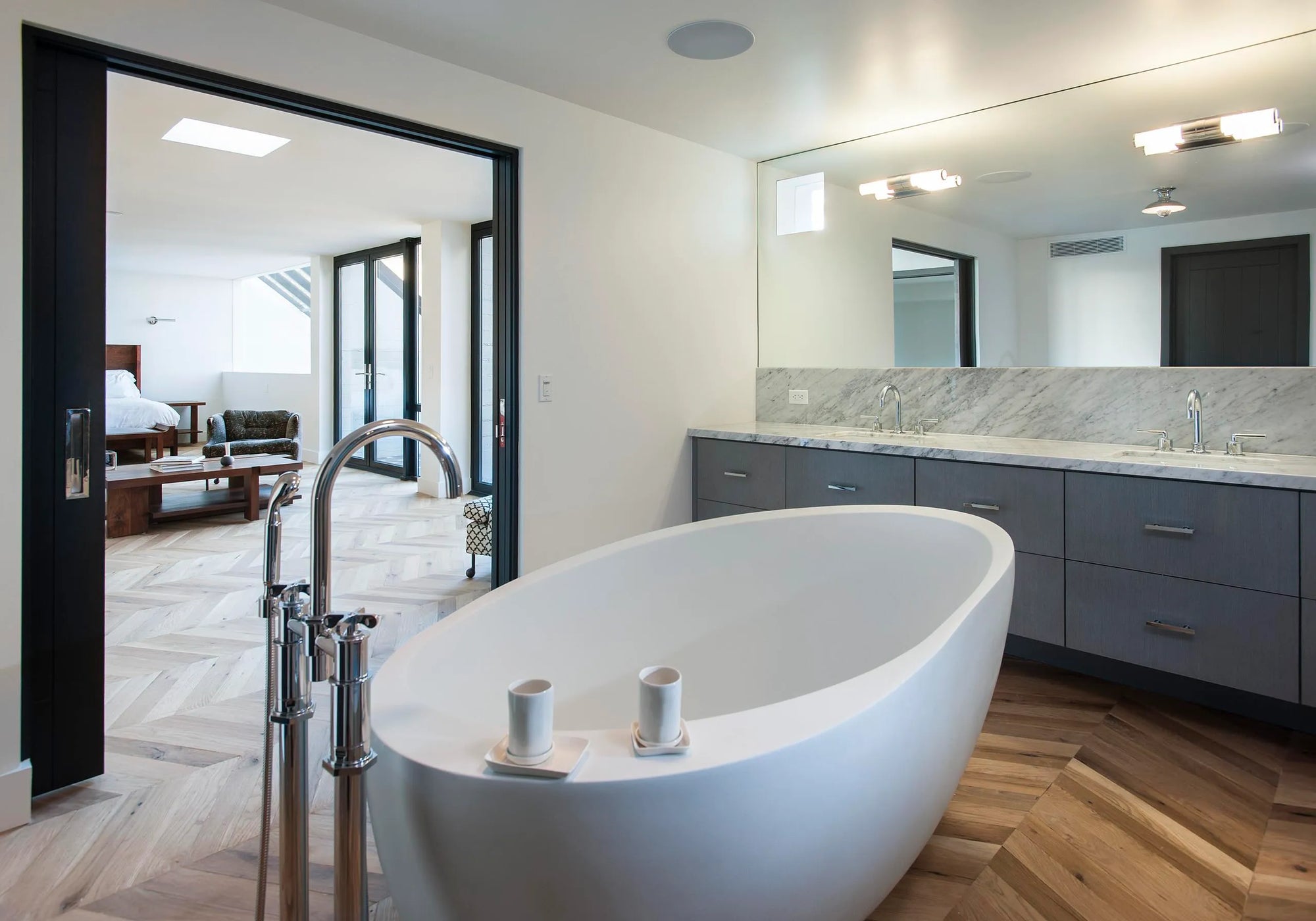 Modern minimalist bathroom with white acrylic bathtub on herringbone wood floor, marble vanity & sleek fixtures.