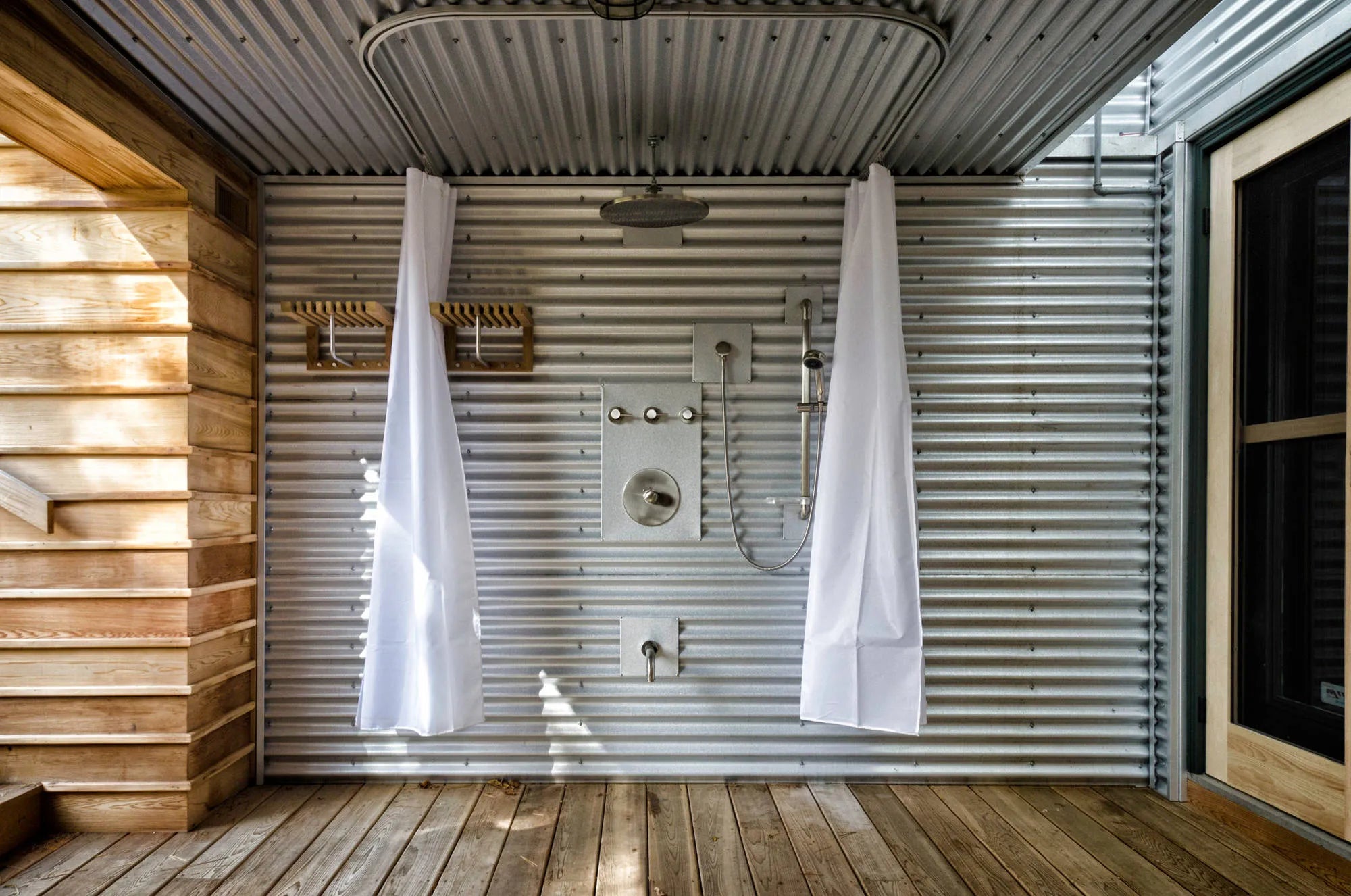 Rustic shower in converted shipping container with corrugated metal walls, wooden accents, and natural light through window.