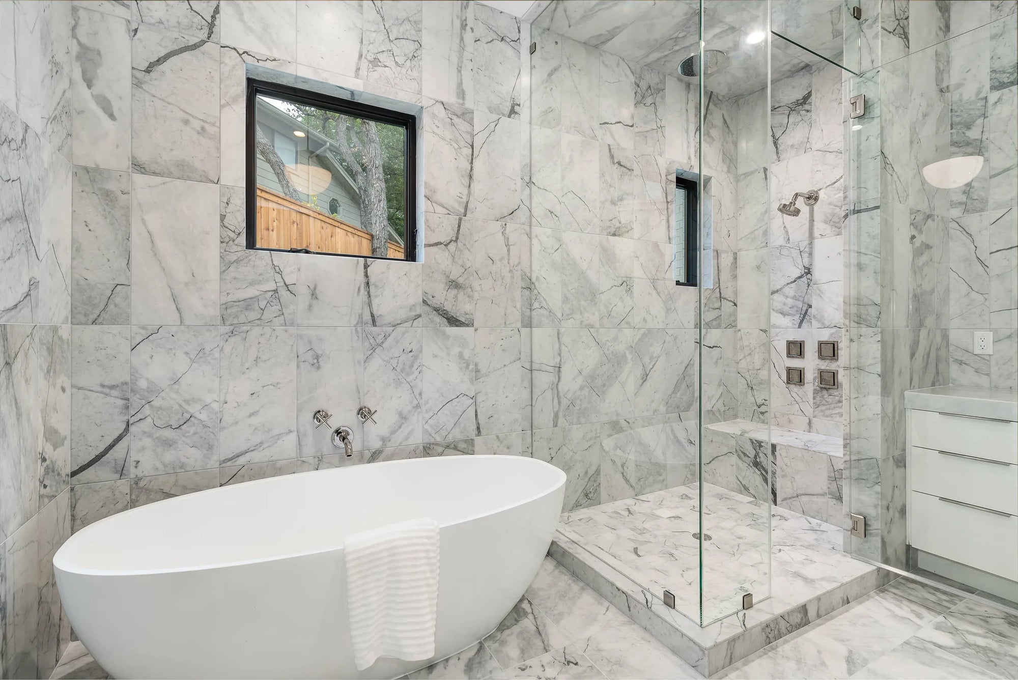 Tub faucet on freestanding bathtub in modern marble bathroom with glass-enclosed shower and natural light.