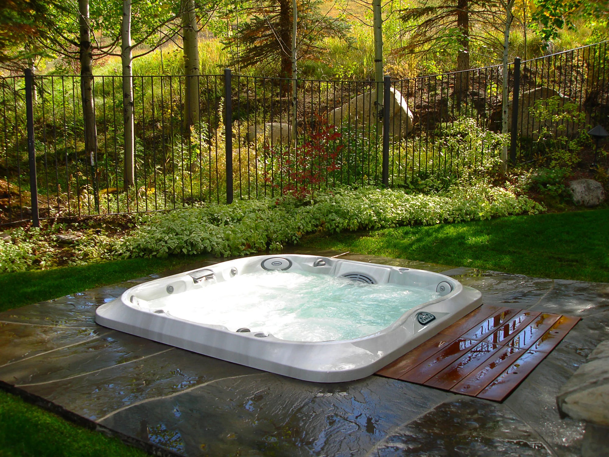 Luxurious outdoor hot tub in a serene backyard setting with bubbling water, lush green grass, and tall trees, surrounded by a black metal fence.