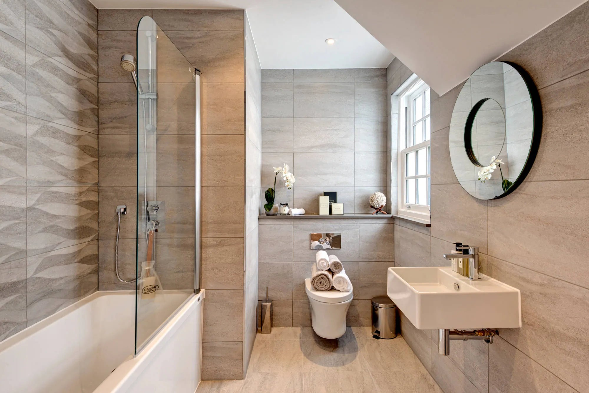 Elegant bathroom with neutral tiles featuring a wall-mounted smart bidet seat, modern sink, round mirror, and glass-enclosed shower-bath combo.
