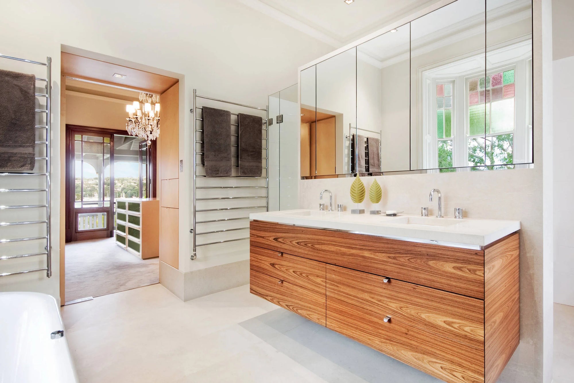 Modern bathroom with wooden vanity, double sinks, large mirror reflecting stained-glass window, and hanging heated towel rack.