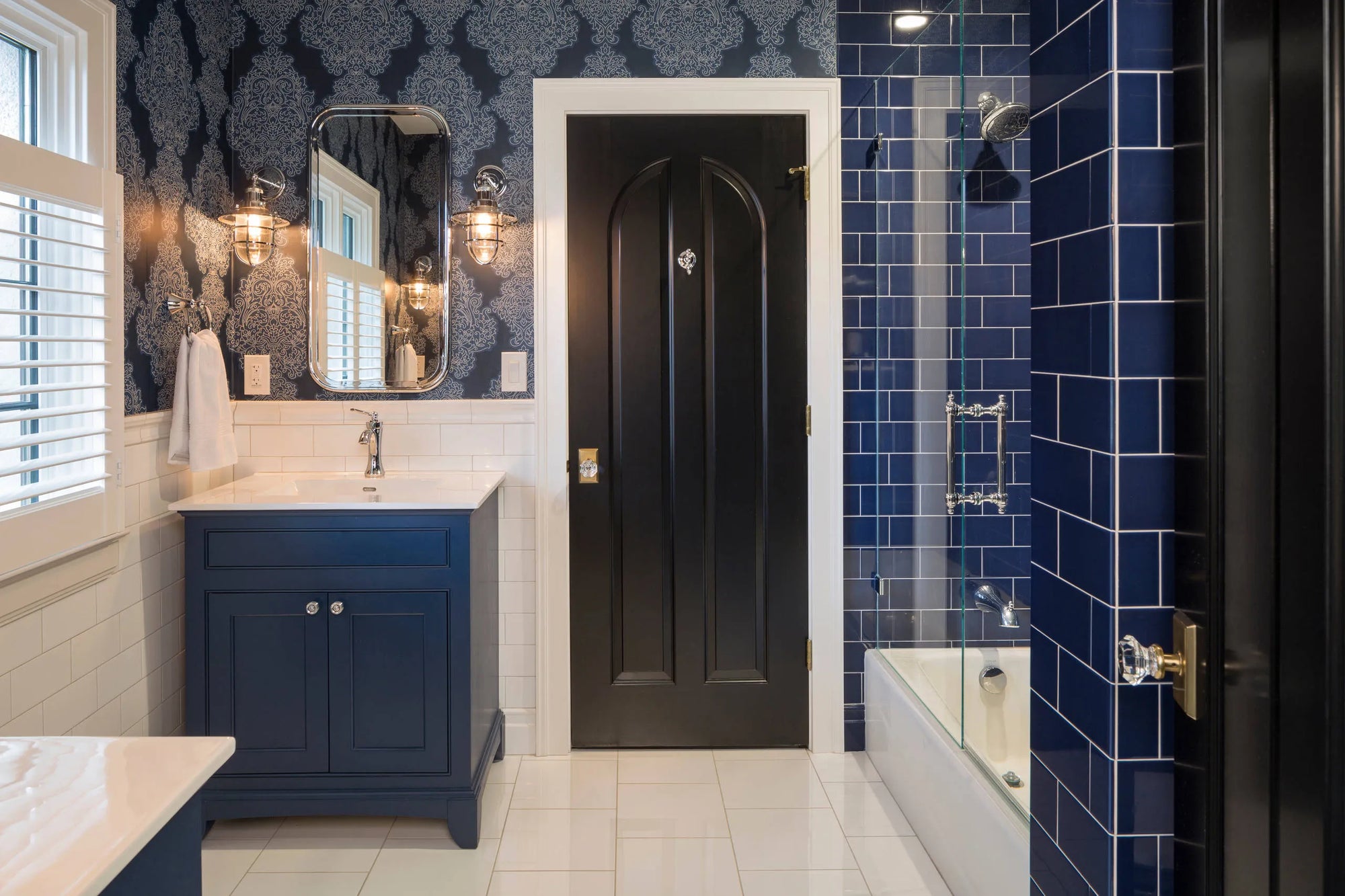 Stylish bathroom with dark door, blue vanity, sink mirror lights, patterned wallpaper, and glass bathtub enclosure amid blue tiles.