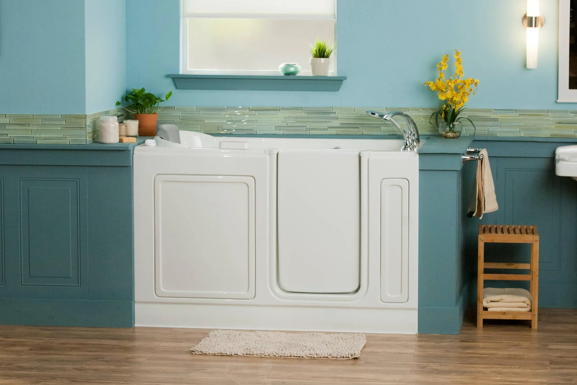Modern white walk-in bathtub with teal blue cabinetry, accessible design, stool, towels, and bath mat.