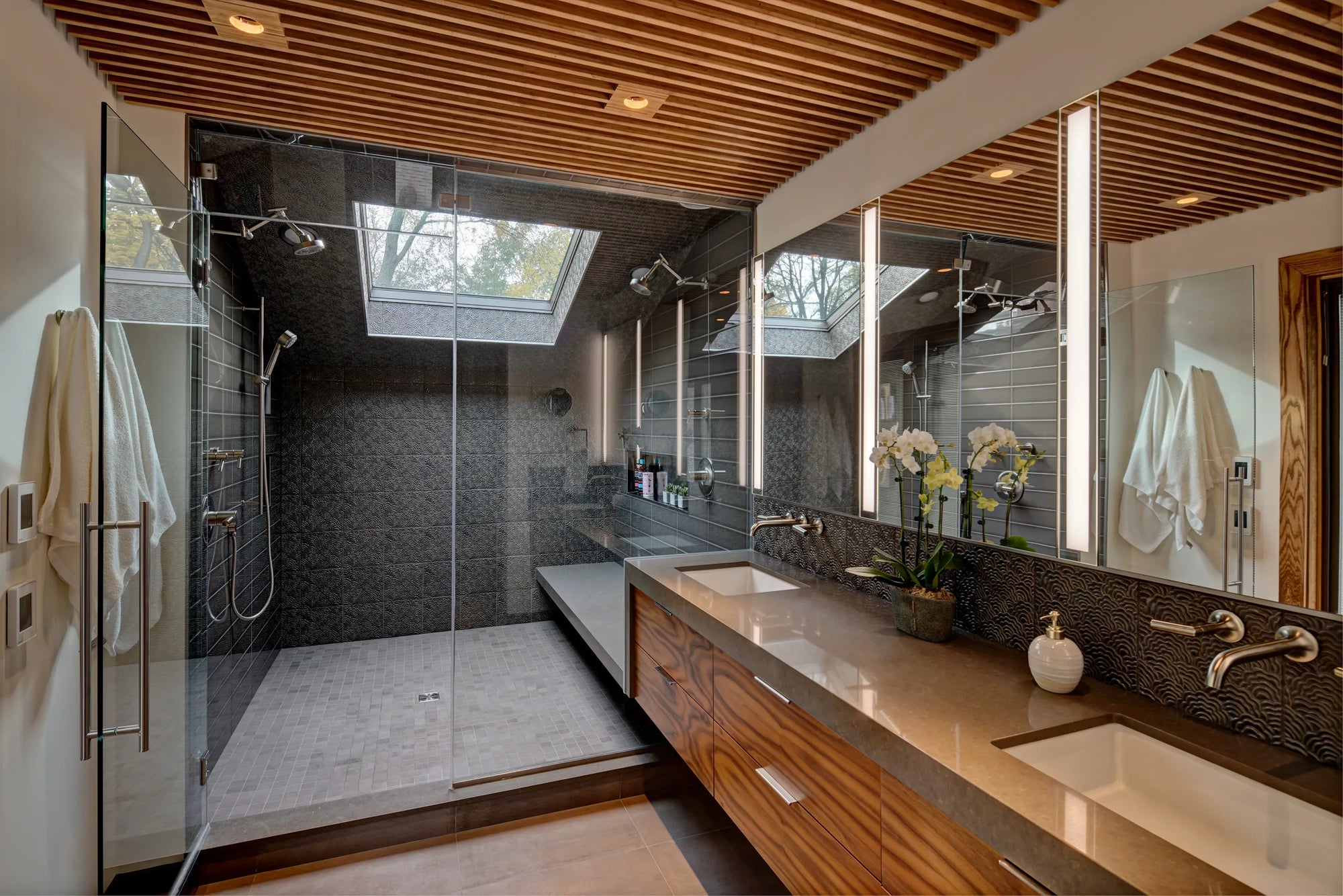 Luxurious modern bathroom with glass walk-in shower and skylight, wooden vanity island, and illuminated mirrors.