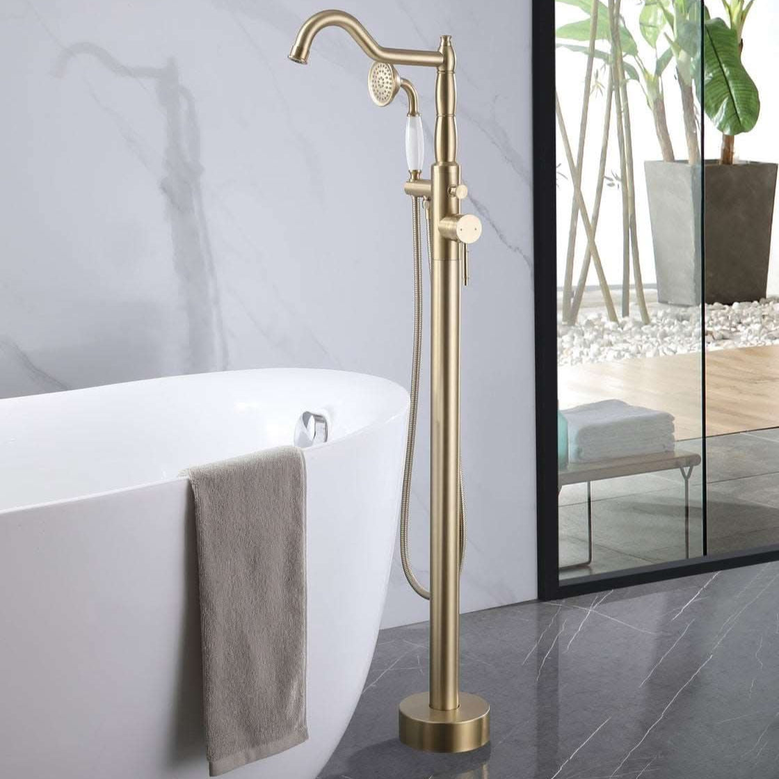 Brushed Gold Freestanding Tub Faucet with Handheld Shower Installed Beside Modern Freestanding White Bathtub.