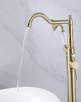 Floor mount brushed gold bathtub filler running water into white soaking tub.