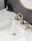 VALMIRA brushed gold sink faucet on white countertop with dark textured wall and gold mirror in background.