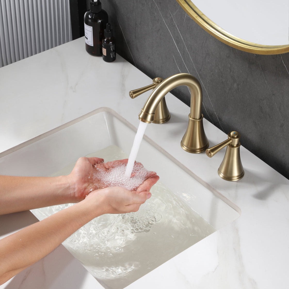 Person washing hands with water from VALMIRA brushed gold sink faucet with curved spout and handles in a modern bathroom.