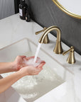 Person washing hands with water from VALMIRA brushed gold sink faucet with curved spout and handles in a modern bathroom.