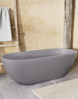 Nectar Series 63-inch matte gray freestanding soaking tub in a bathroom with beige walls, wooden beam, towel rack, and gray stone tile.