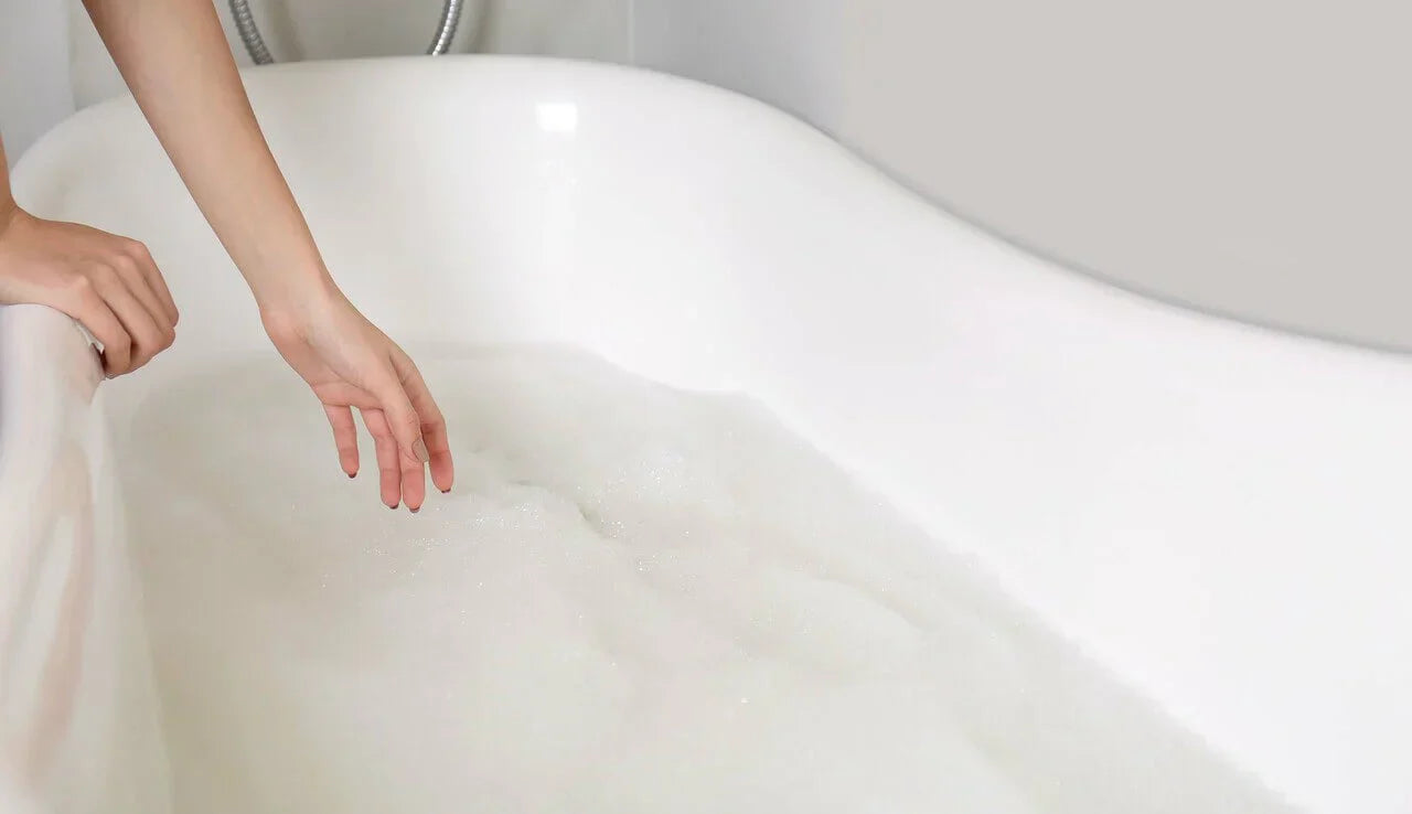 Close-up of hands on a pristine white bathtub with fluffy bubbles, evoking luxury relaxation in a minimalist bathroom.