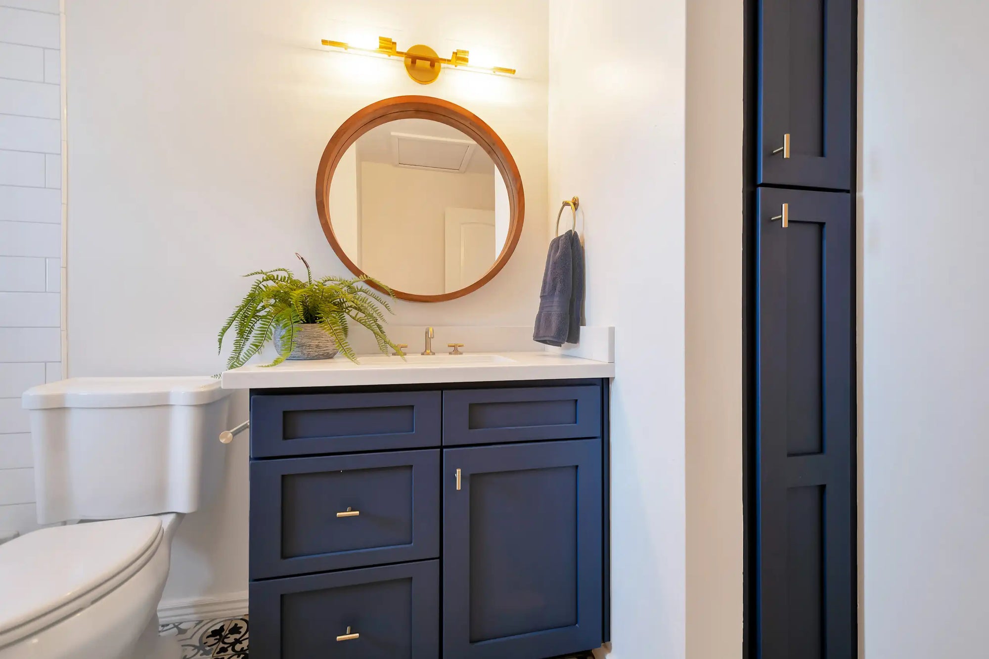 A clean, modern bathroom featuring a navy blue freestanding single bathroom vanity with brass hardware and a round mirror accent. | ACE DECOR