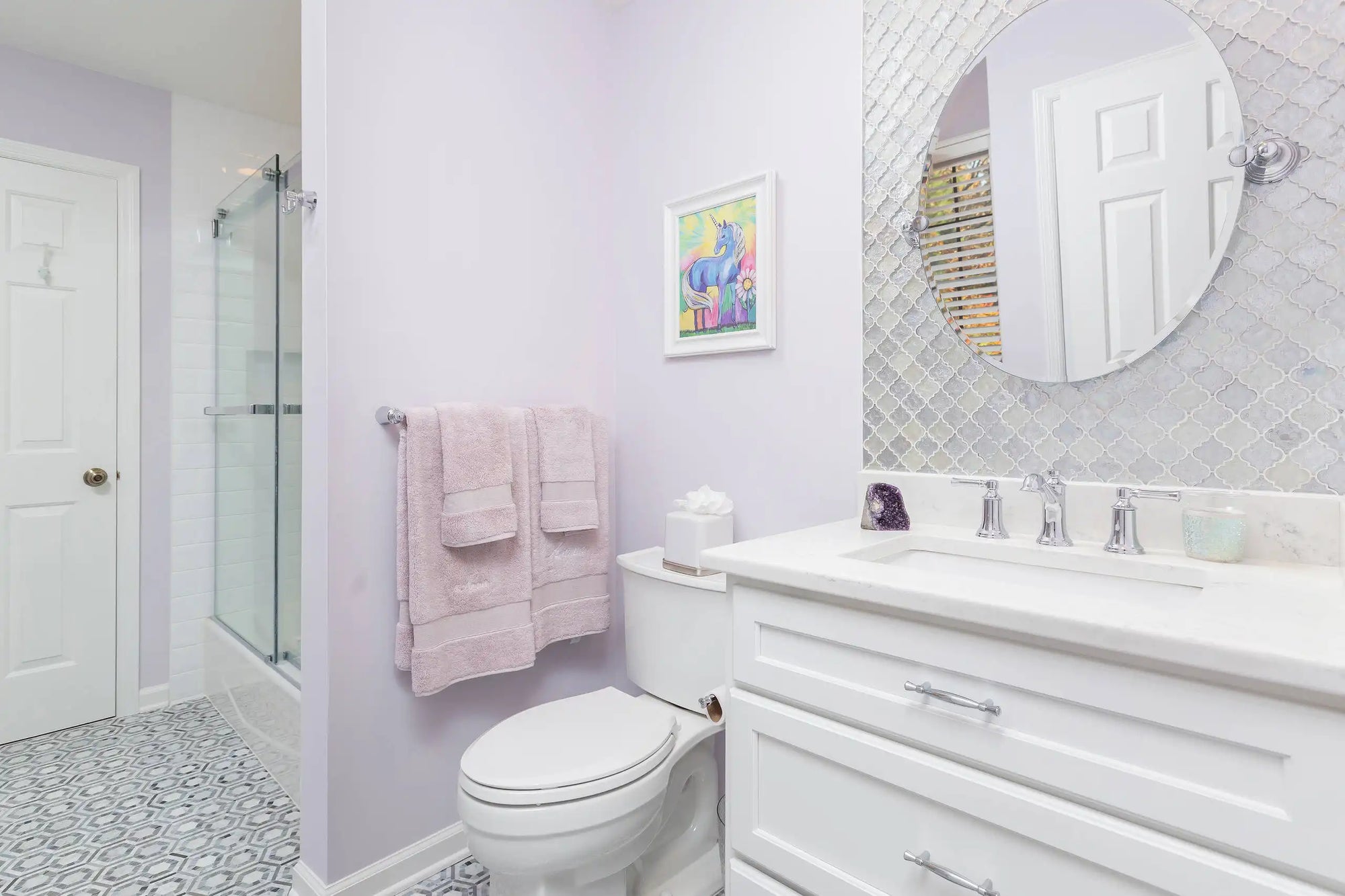 A bright bathroom with a white vanity, oval mirror, glass shower, and pastel purple accents. |ACE DECOR