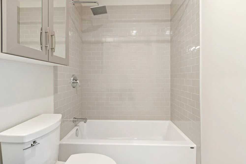 a modern alcove bathtub with light gray subway tile walls, a square rainfall showerhead, and a sleek built-in cabinet above the toilet. | ACE DECOR