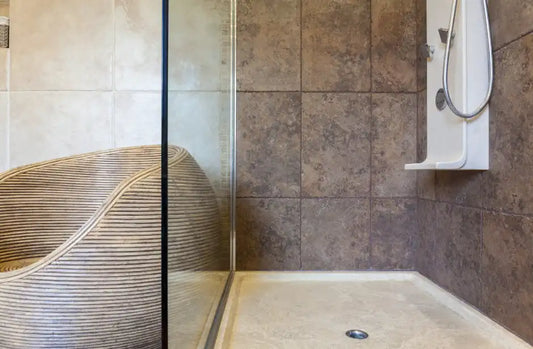 A close-up photo of the corner of an elegant, modern bathroom with a curved glass door, tile walls, and a wicker chair in front of it. The design is modern, with natural stone tiles on the wall and a beige and brown color scheme. | ACE DECOR