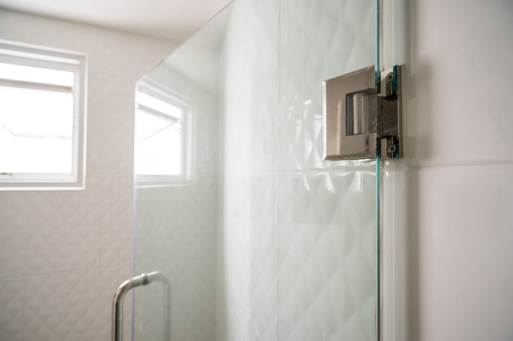 A close-up view of a frameless glass shower door with a metal hinge and handle in a modern tiled bathroom. | ACE DECOR