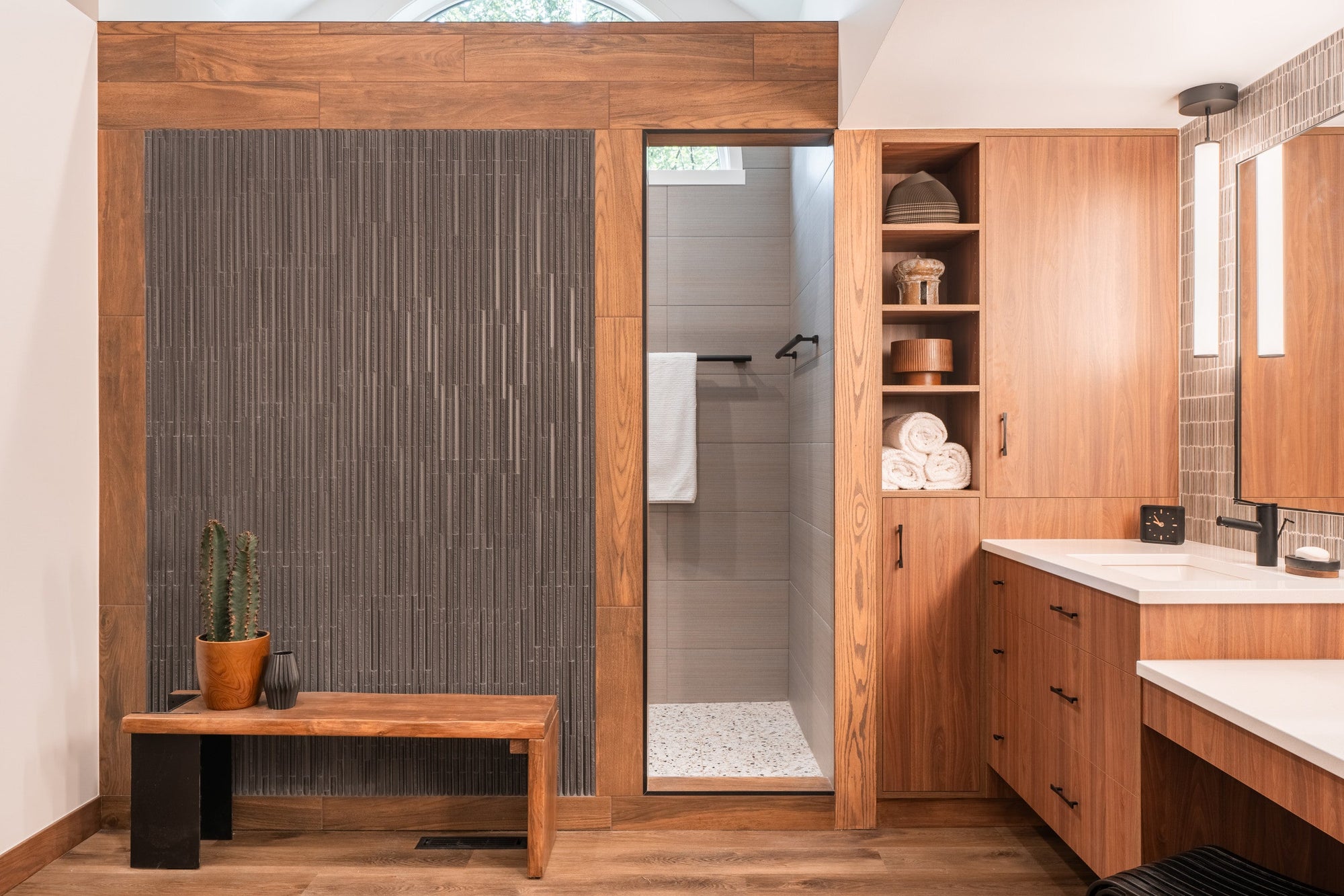 A modern, spa-inspired bathroom featuring warm wood cabinetry, textured black tile, and a sleek black-framed shower entryway. | ACE DECOR