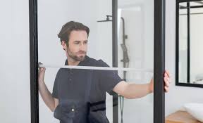 A man is measuring a frameless shower door panel with a tape measure during installation. | ACE DECOR