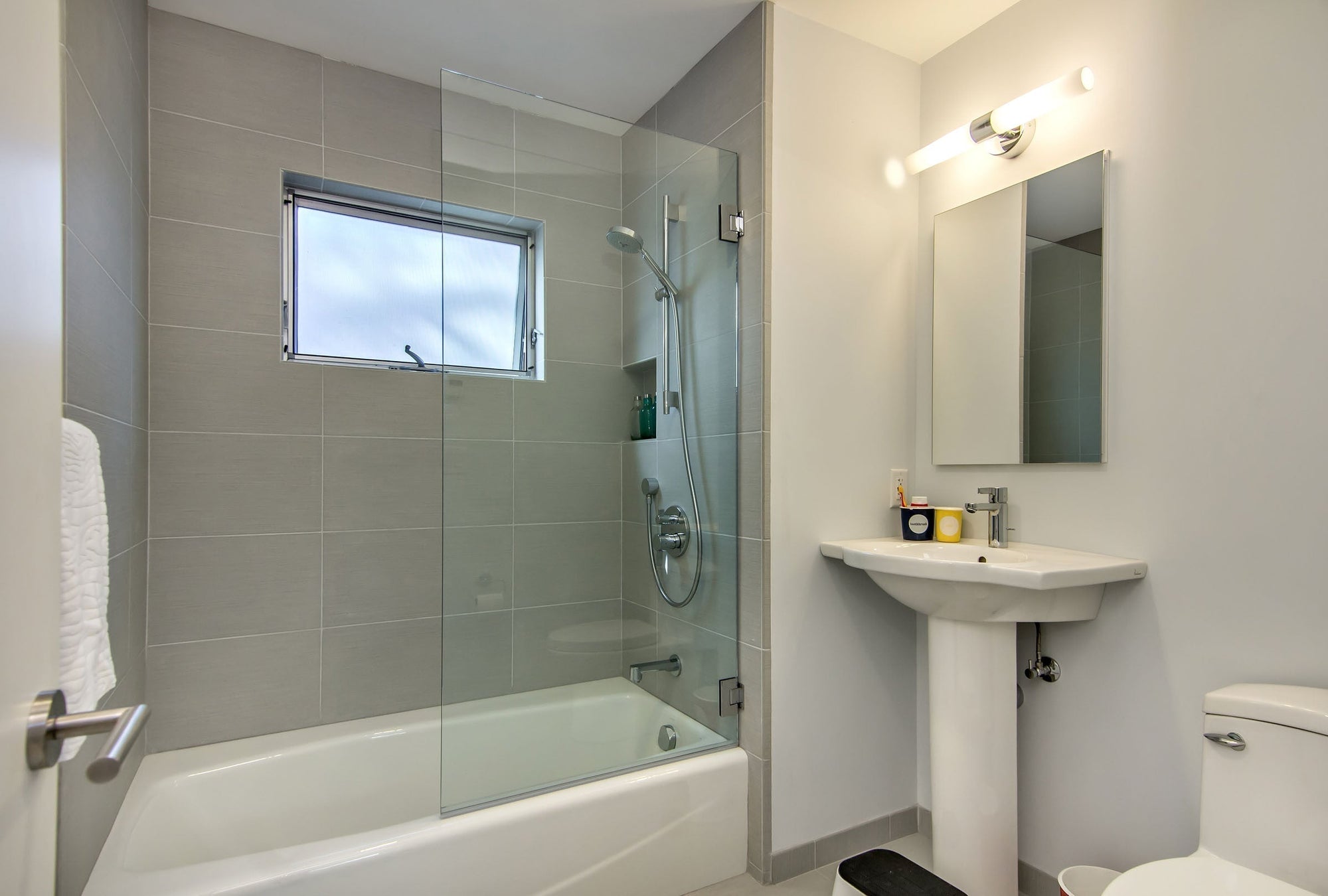 A clean and minimalist bathroom featuring a half glass shower door on the bathtub, complemented by neutral gray tiles, a pedestal sink, and a modern toilet. |ACE DECOR