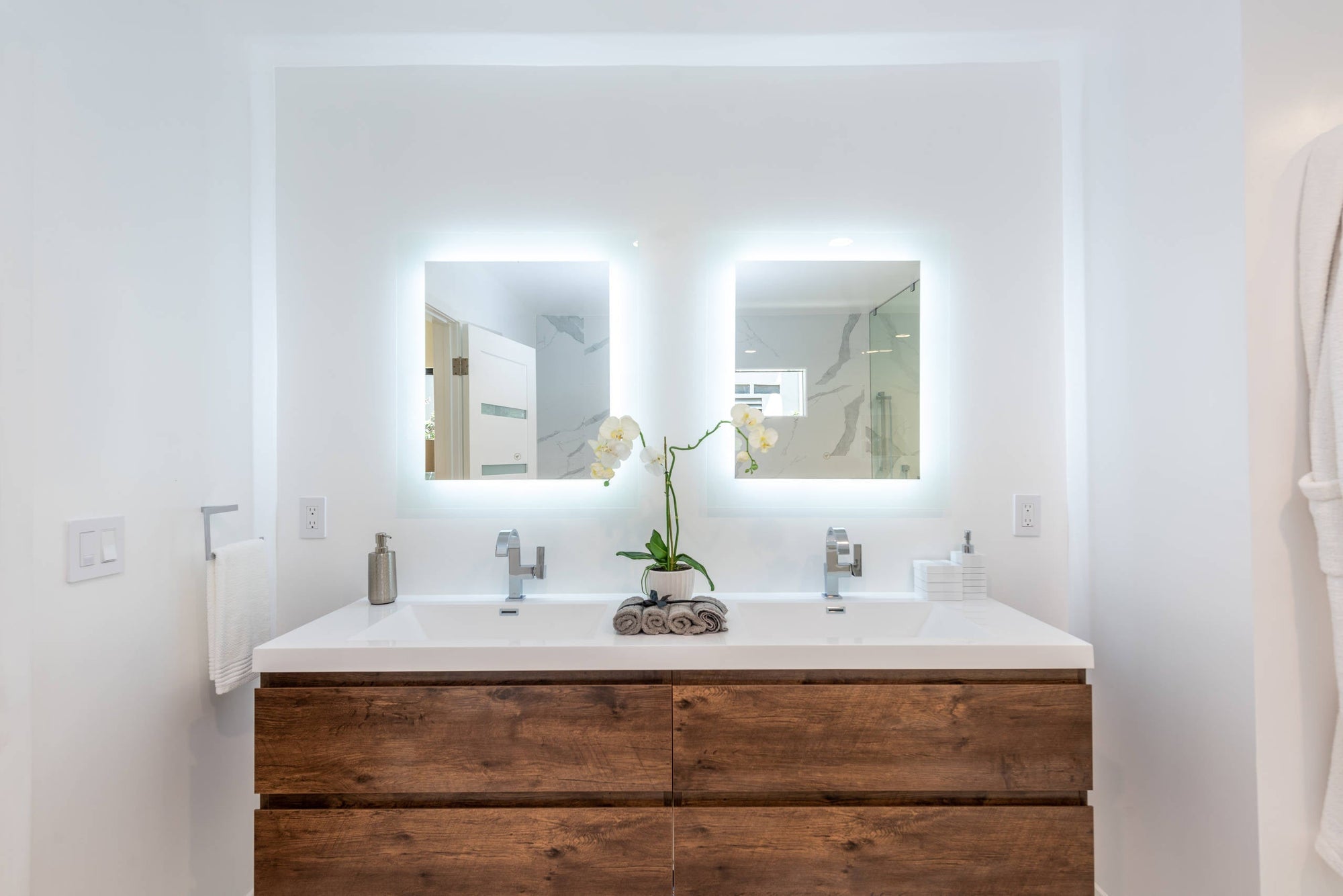 A clean, modern bathroom setup featuring two square LED mirrors above a double sink vanity with natural wood drawers. | ACE DECOR