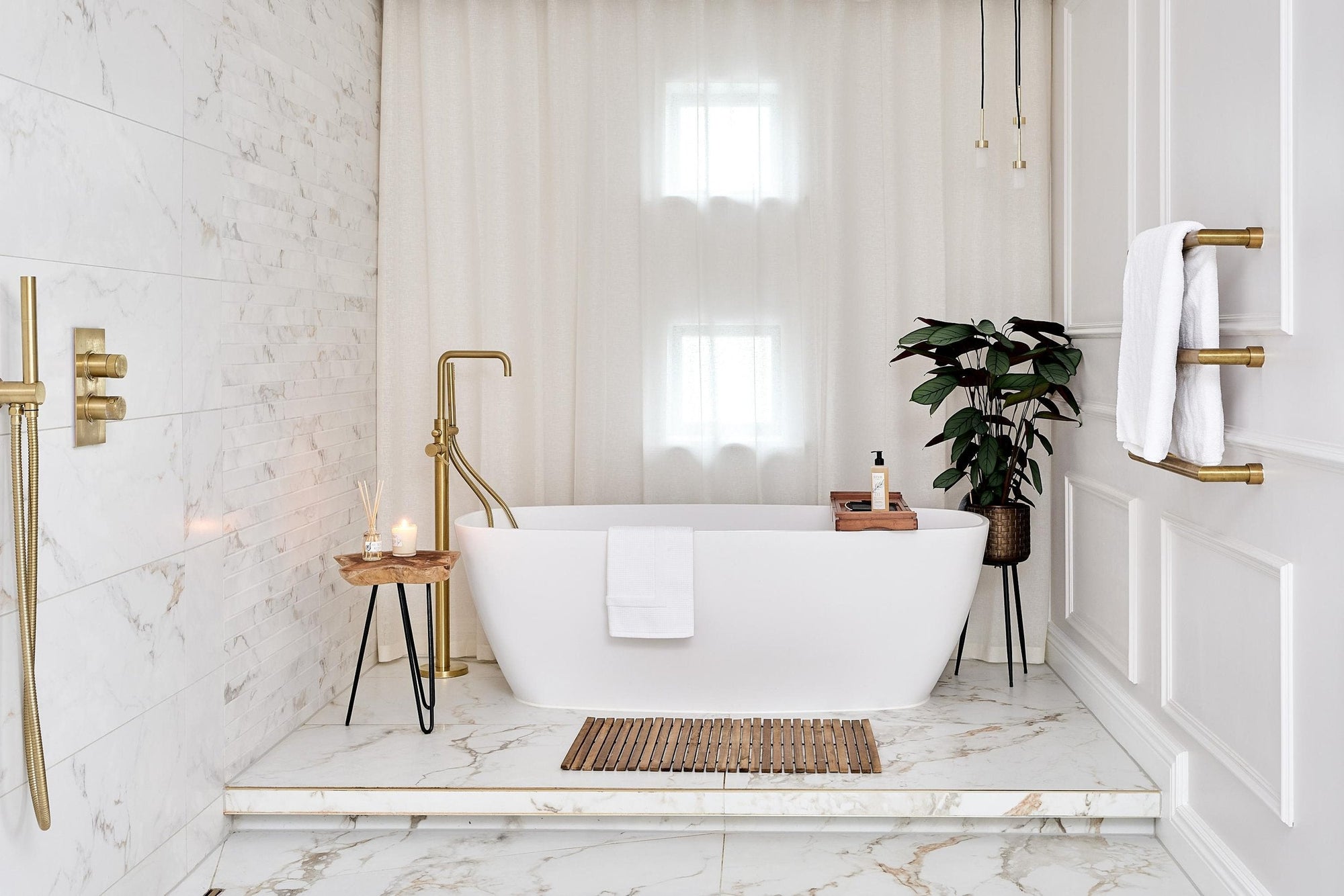 a stylish bathroom with a freestanding bathtub, marble floors, a wooden bath mat, and a touch of greenery, all accented by gold fixtures. | ACE DECOR