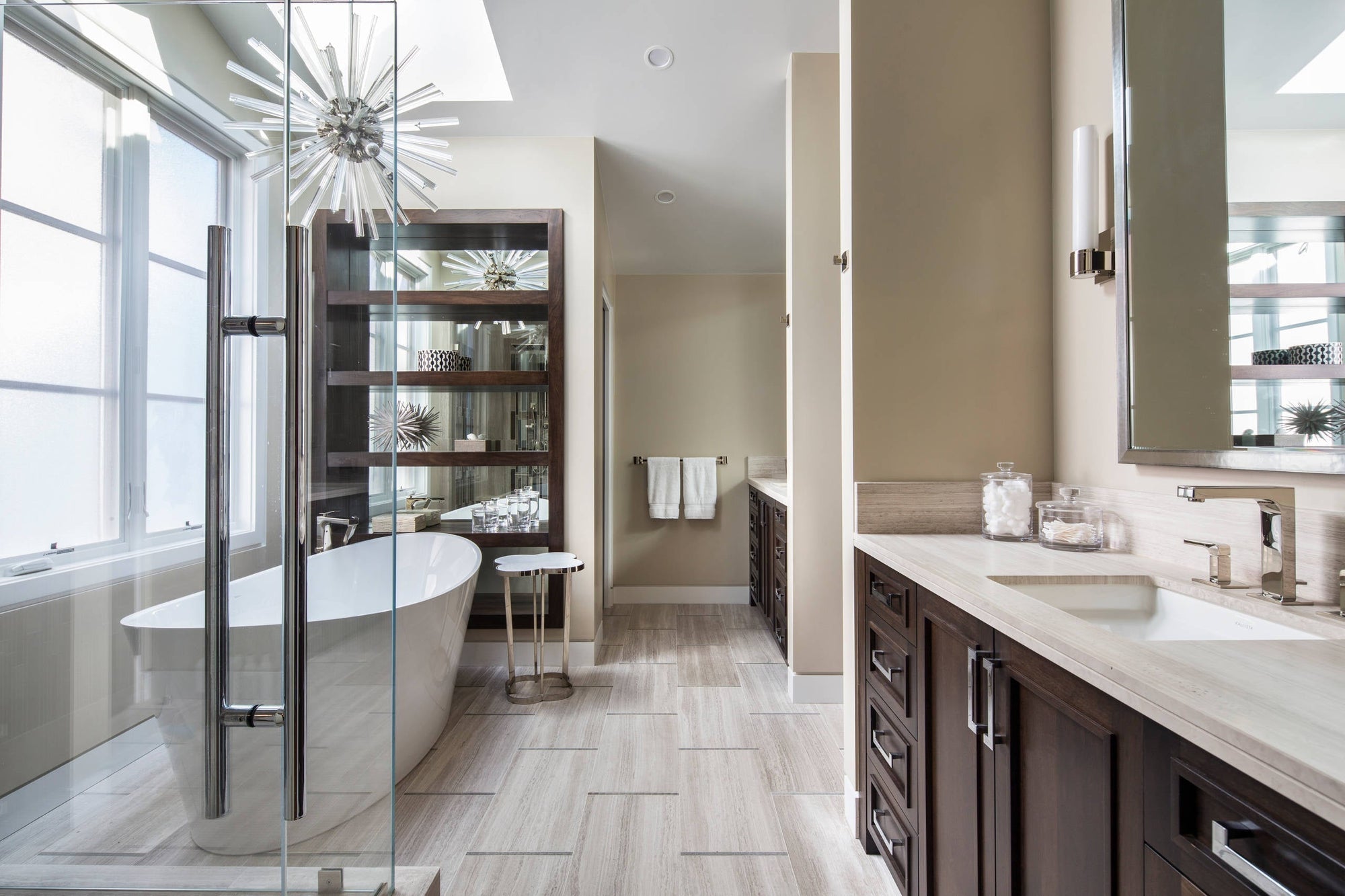 A modern minimalist bathroom features a round freestanding bathtub, floor-to-ceiling windows flooding natural light, light gray hexagonal tiles, dual marble sinks with brushed gold fixtures, and a glass-walled rainfall shower.
