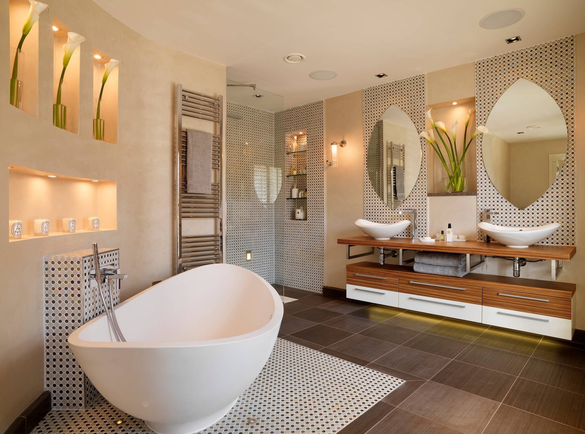 A luxurious modern bathroom featuring a freestanding marble bathtub, dual vanities with mirrored cabinets, lush potted plants, and neutral tones accented by warm lighting in an open-plan design.
