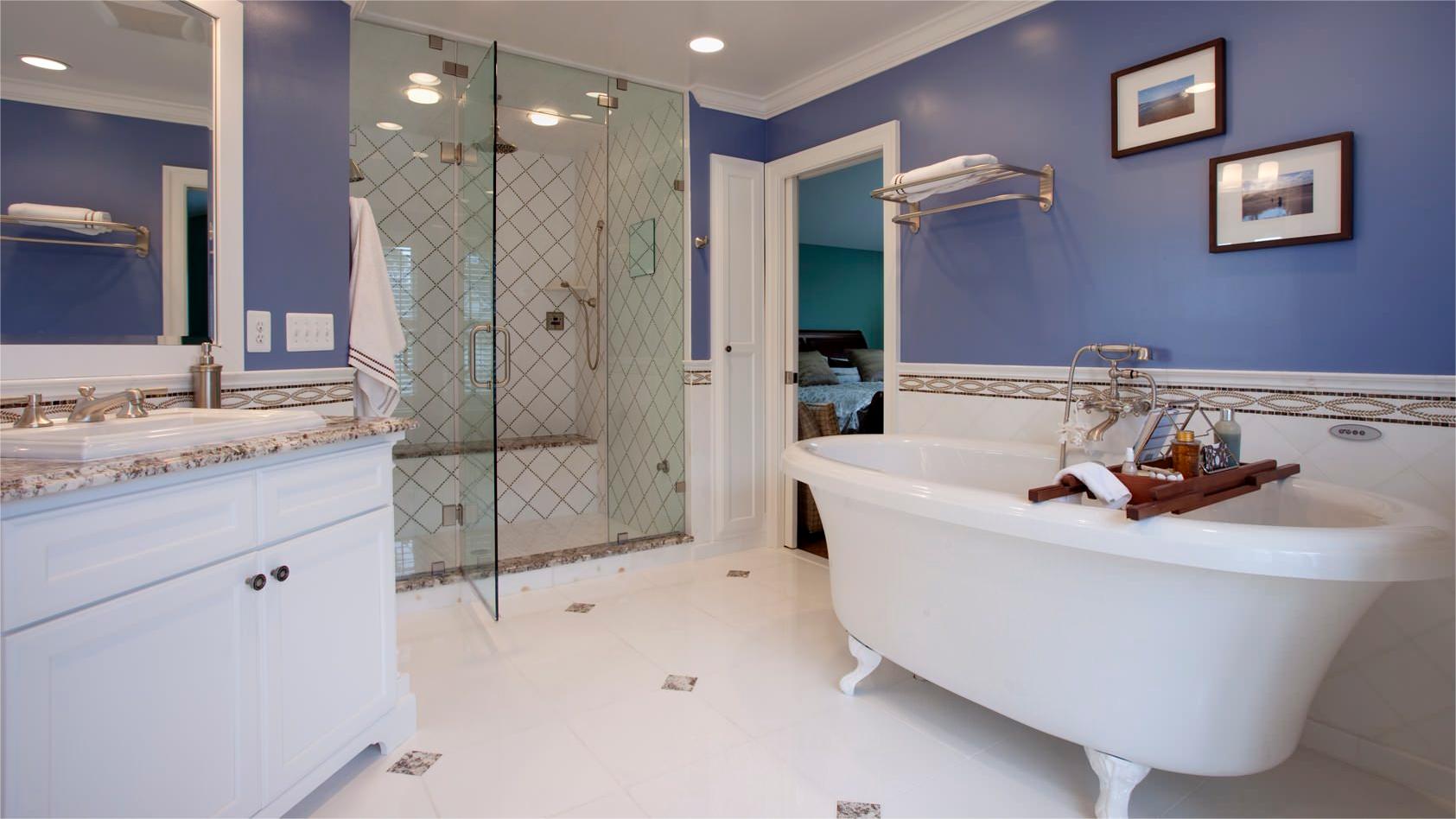A serene blue bathroom with a clawfoot bathtub, glass-enclosed diamond-patterned shower, white marble vanity with granite tops, decorative trim, framed art, and tiled flooring blending modern/traditional elegance.