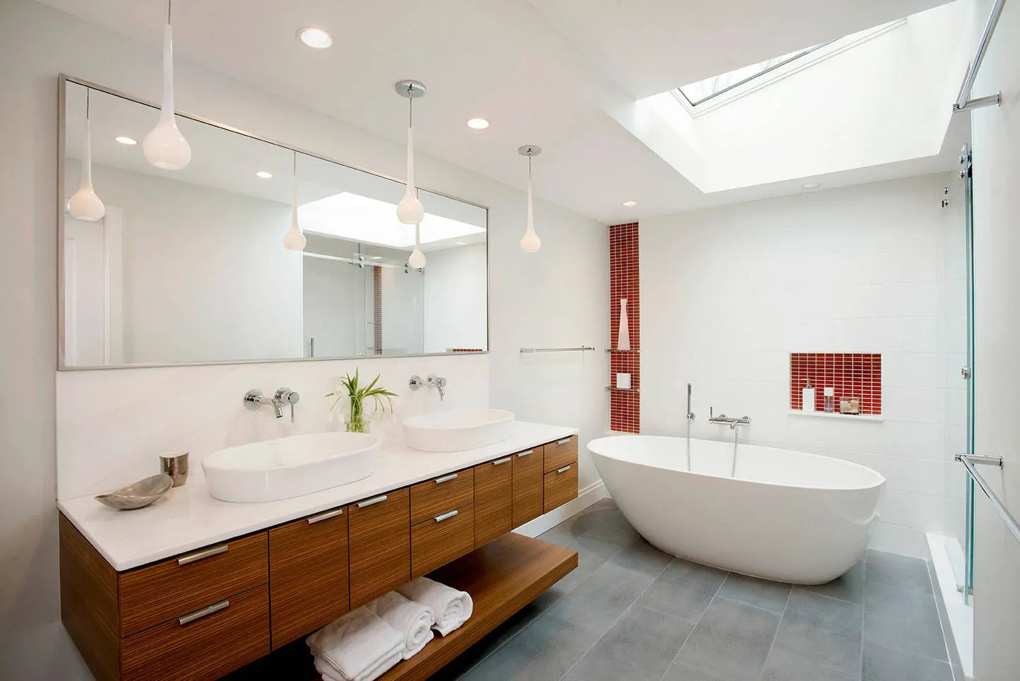 Modern bathroom with wooden vanity sinks, pendant lights, white freestanding bathtub, gray flooring, and red-tiled accent wall under natural skylight light.