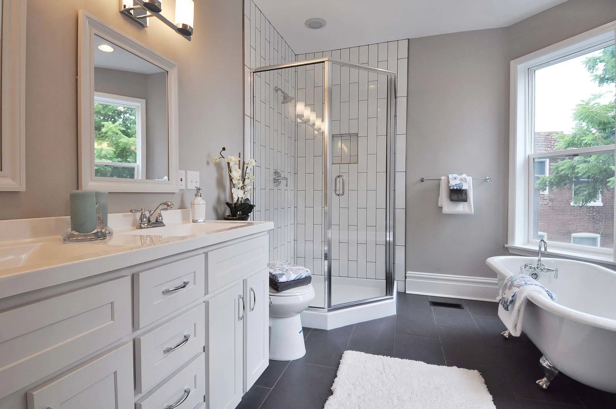 A modern bathroom with walk-in shower, double vanity, freestanding tub, light gray walls, dark wood floors, and abundant natural light.