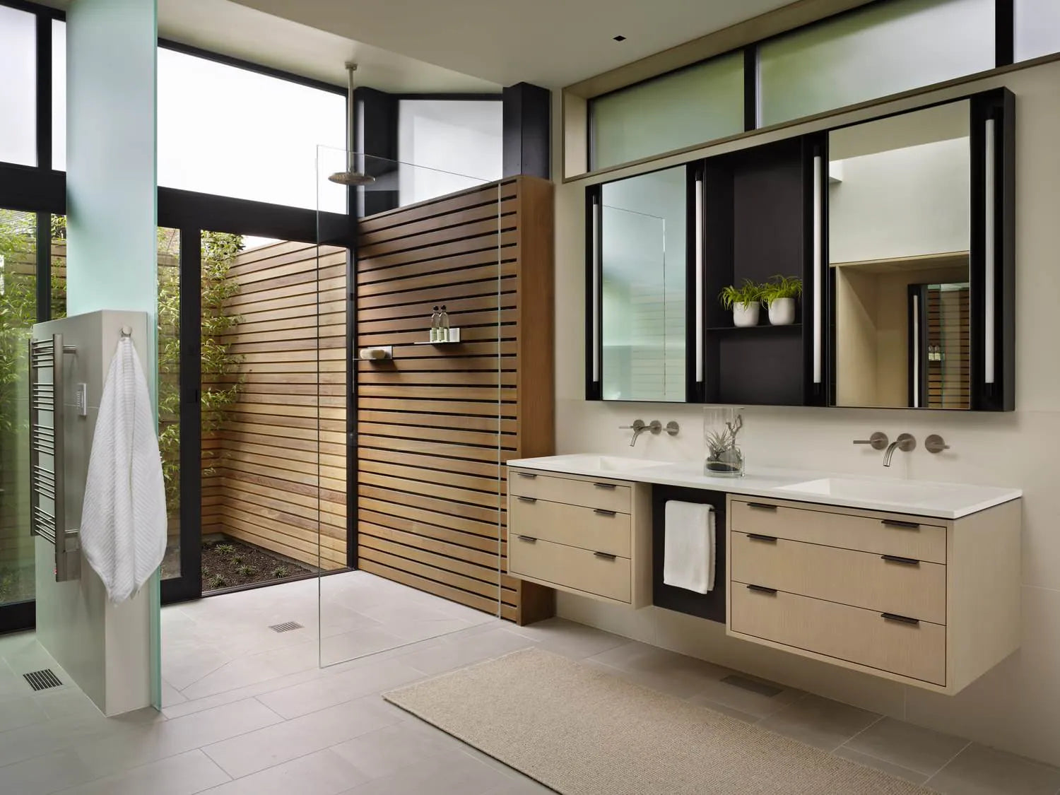 Modern bathroom with double vanity, glass shower, natural light, and neutral tones, featuring wooden accents and greenery views.