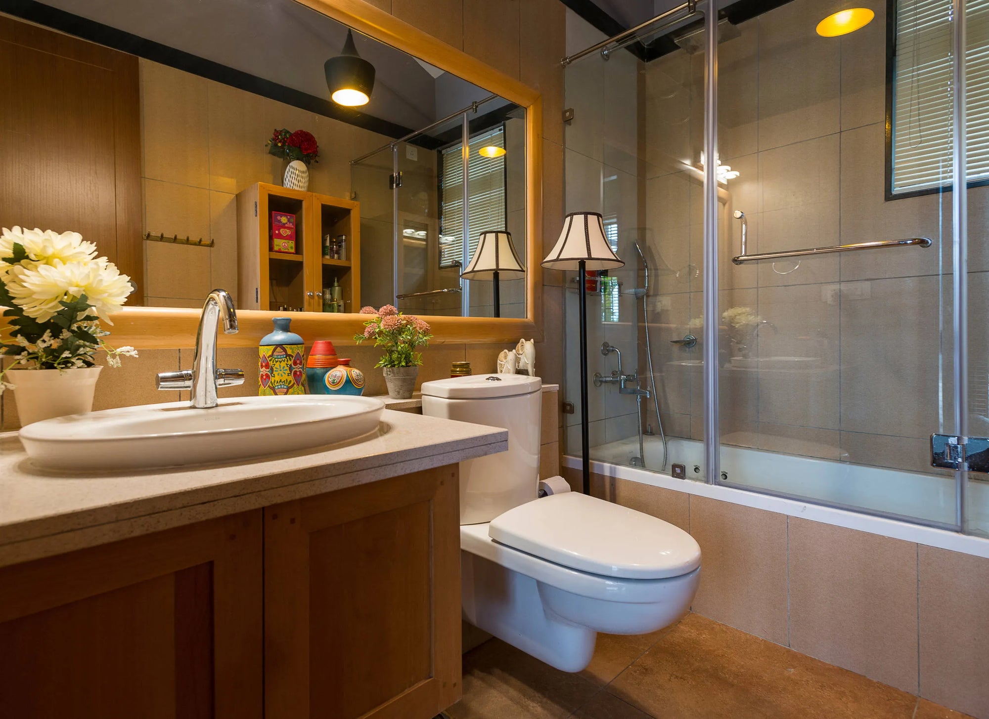 Modern bathroom featuring glass bathtub enclosure, white sink with wooden vanity, large mirror, and decorative accents including fresh flowers under warm ambient lighting.