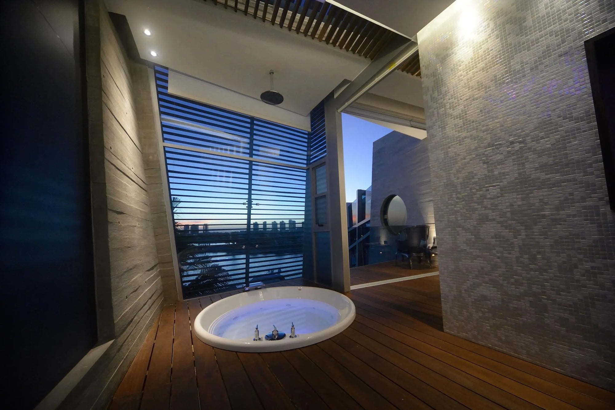 Modern in-floor bathtub with glowing blue water, wooden floors, tiled walls, and large windows overlooking twilight scenery.