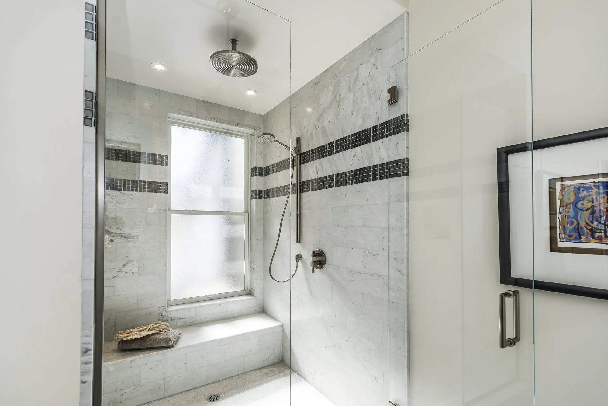 Modern bathroom shower with marble-look tiles, natural light window, wooden bench, and framed wall artwork in contemporary design.