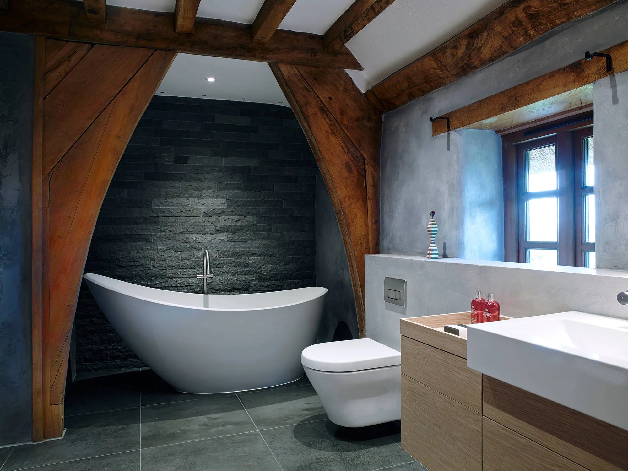 Modern bathroom featuring white wall hung smart toilet beside freestanding bathtub, dark stone walls, wood vanity, and natural light.​