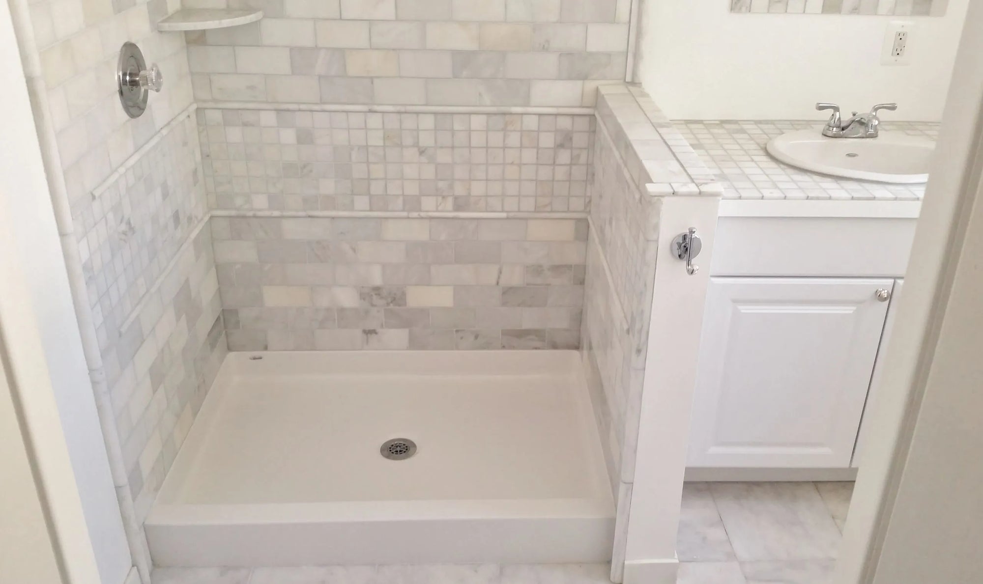 White square shower base with centered drain and level-entry design, adjoined by vanity, with light gray mosaic tile walls in neutral modern bathroom.​