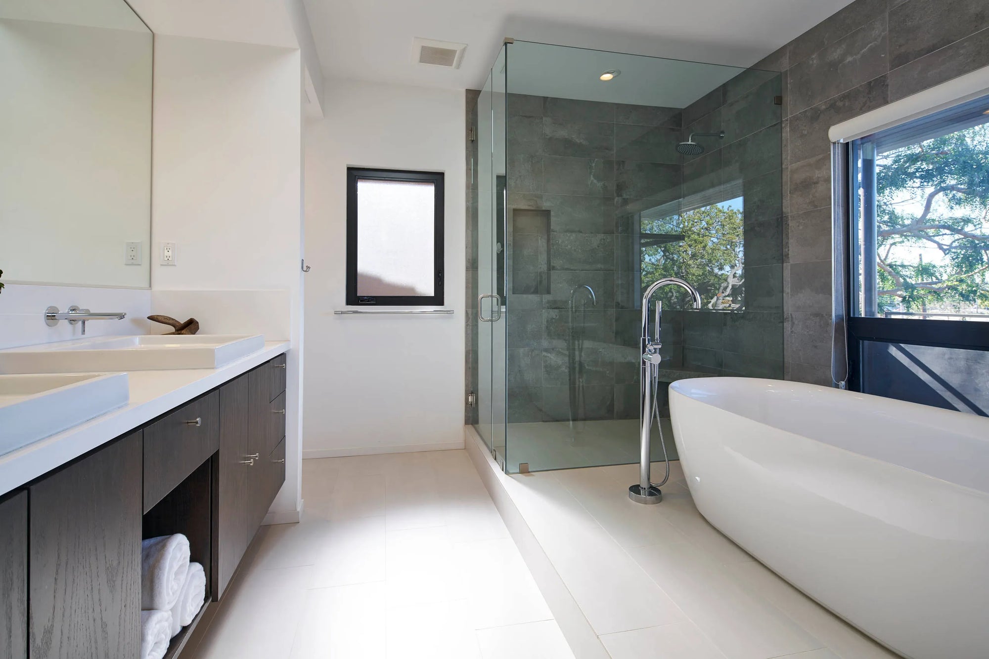 Modern bathroom with freestanding bathtub near window, glass shower, dark wood vanity, and bathtub faucets with spray hoses.​