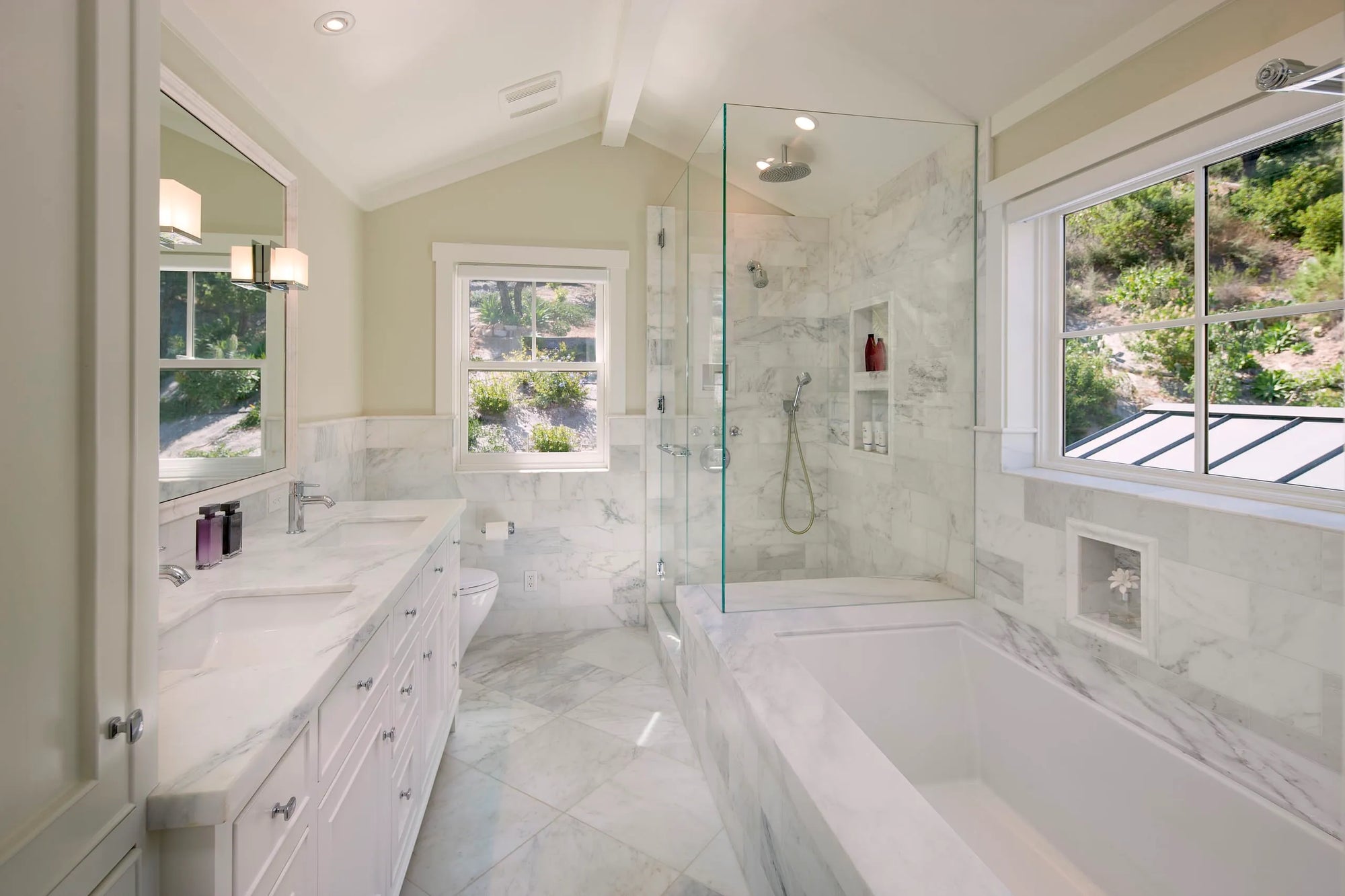Elegant white bathroom with glass shower door, double vanity, marble tiles, freestanding tub, and large greenery-view windows.